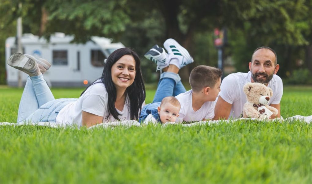 Happy family lying on grass with RV in the background, enjoying a sunny outdoor picnic.
