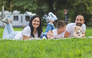 Happy family lying on grass with RV in the background, enjoying a sunny outdoor picnic.