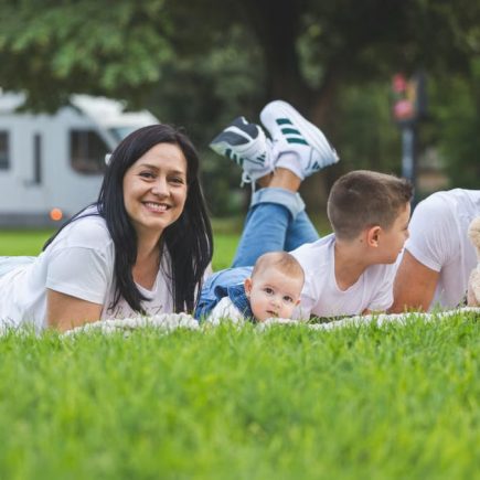 Happy family lying on grass with RV in the background, enjoying a sunny outdoor picnic.